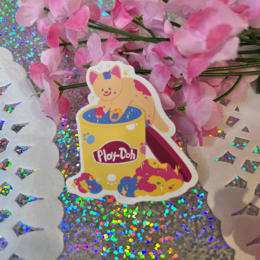 Sticker of the Nostalgic Clay Cat (v.3), depicting a professionally made white cartoon cat playing in a yellow Play-Doh container with multicolored dough on its paws and surrounding area. The background features a sparkly holographic surface and delicate pink flowers. Crafted from waterproof matte vinyl, it's both adorable and durable.