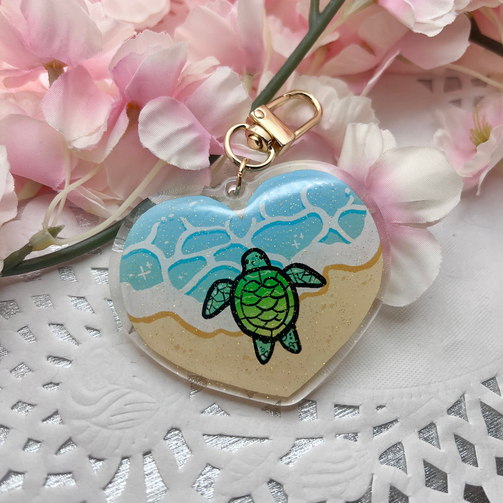 A keychain charm that is heart shaped, showing the ocean tide on sand, with a turtle in the middle