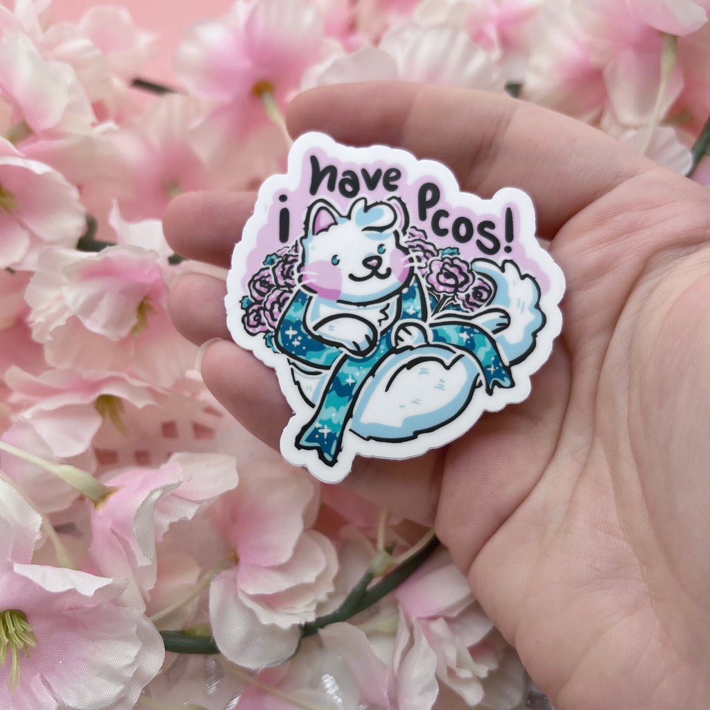 a sticker of a white cat surrounded by pink carnations. the cat is holding a teal condition ribbon and above them is the text "i have pcos"