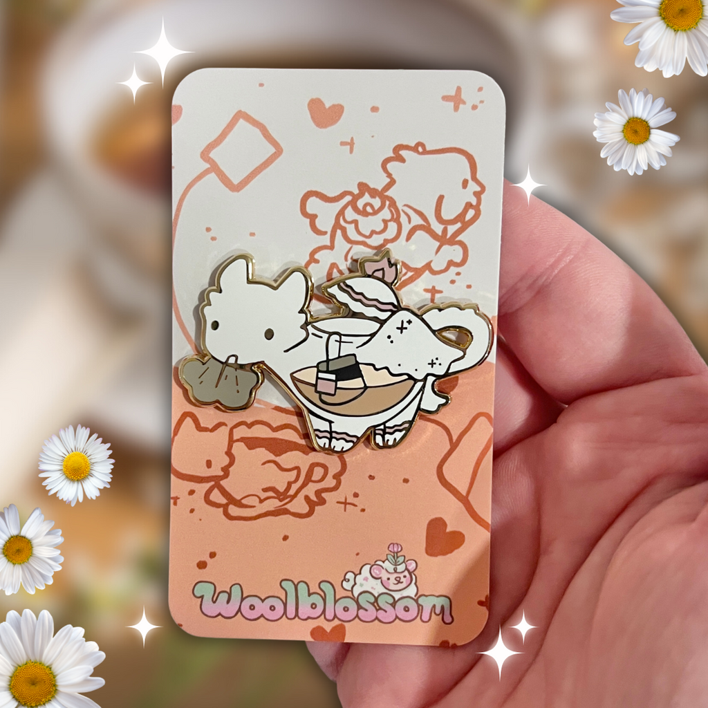 Card with cartoon character dragon pin and 'Woolblossom' branding held by a hand, surrounded by daisies.