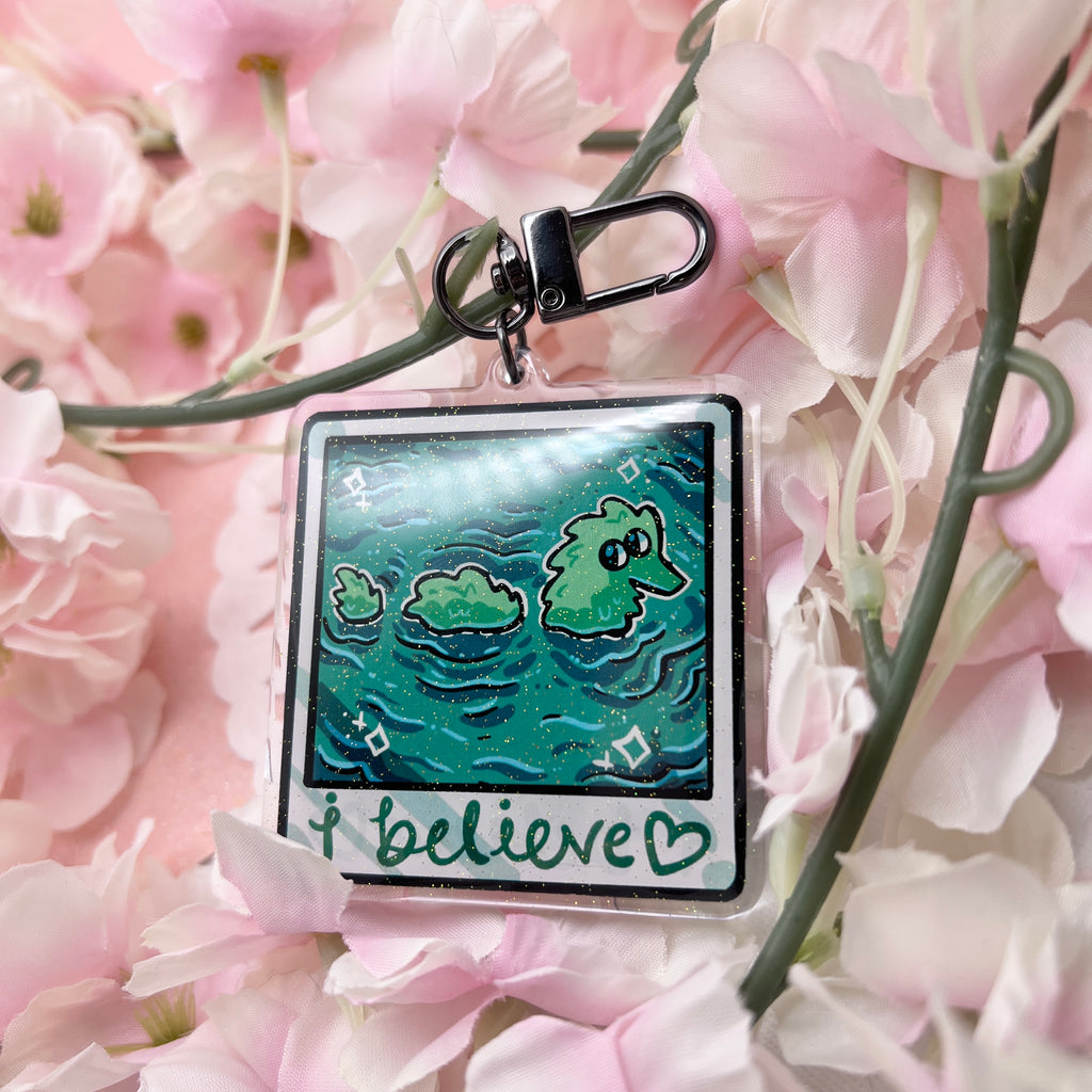 A keychain that looks like a camera photo, hand written text at the bottom that says "i believe <3", in the photo is lake water with a fuzzy worm lochness monster