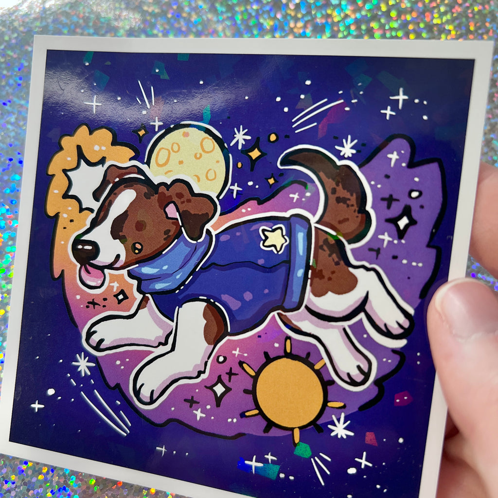 Print of a dog in a space-themed outfit on a glittery background held by hand