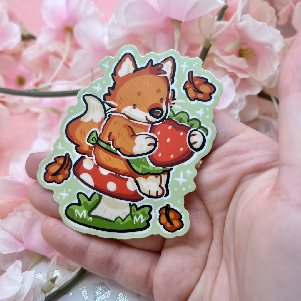 A sticker of a red fox with a little green apron sitting on a red mushroom, holding a strawberry with autumn leaves falling