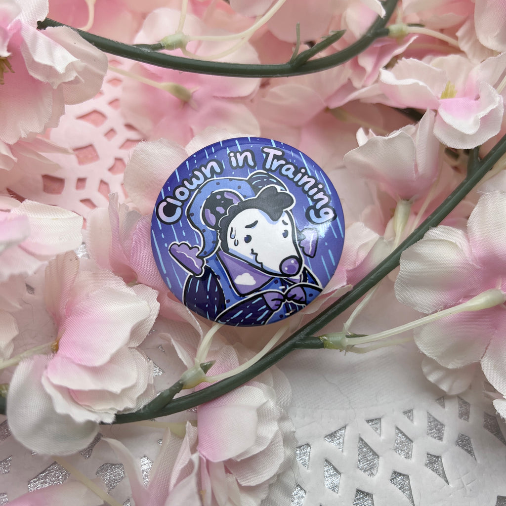 A button of a rain themed opossum that looks a little nervous with the text "clown in training"