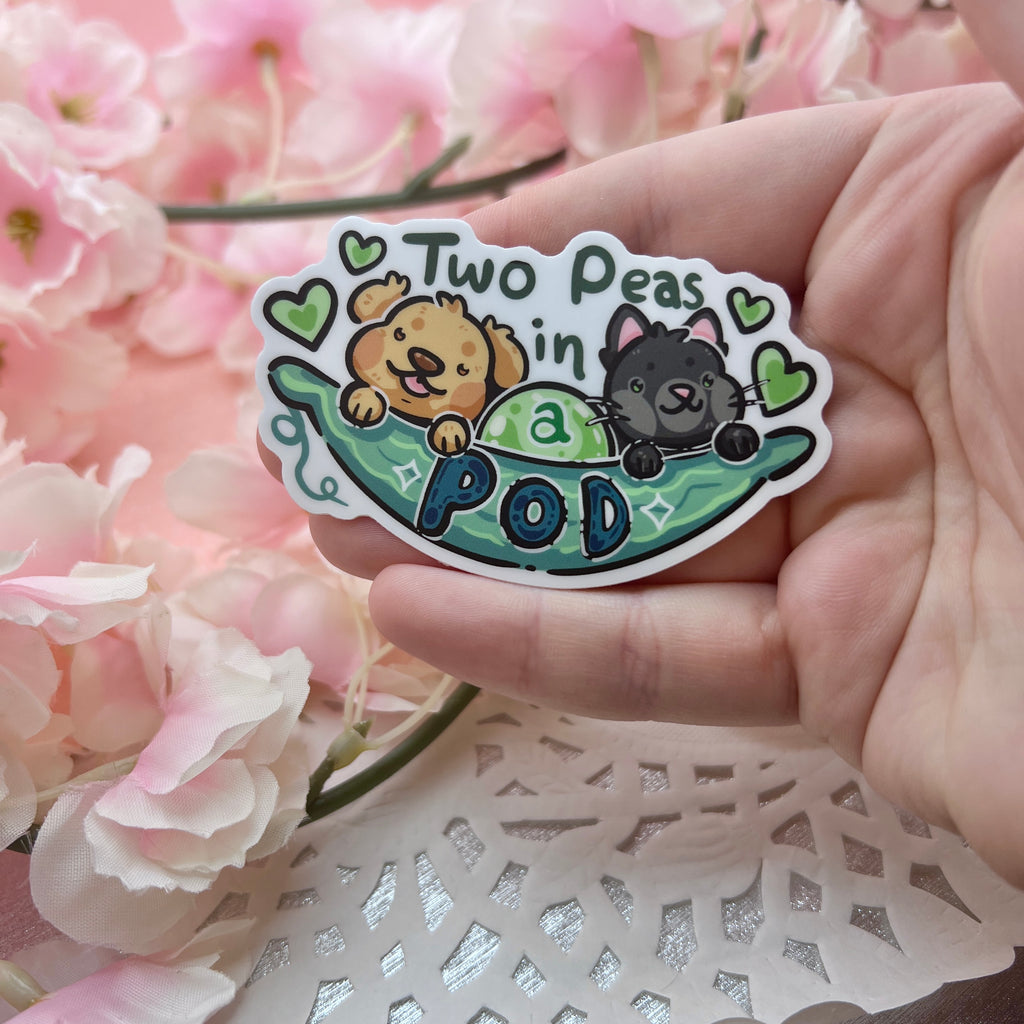 A sticker of a pea pod with a golden retriever and a black cat sticking out and text that says “two peas in a pod”