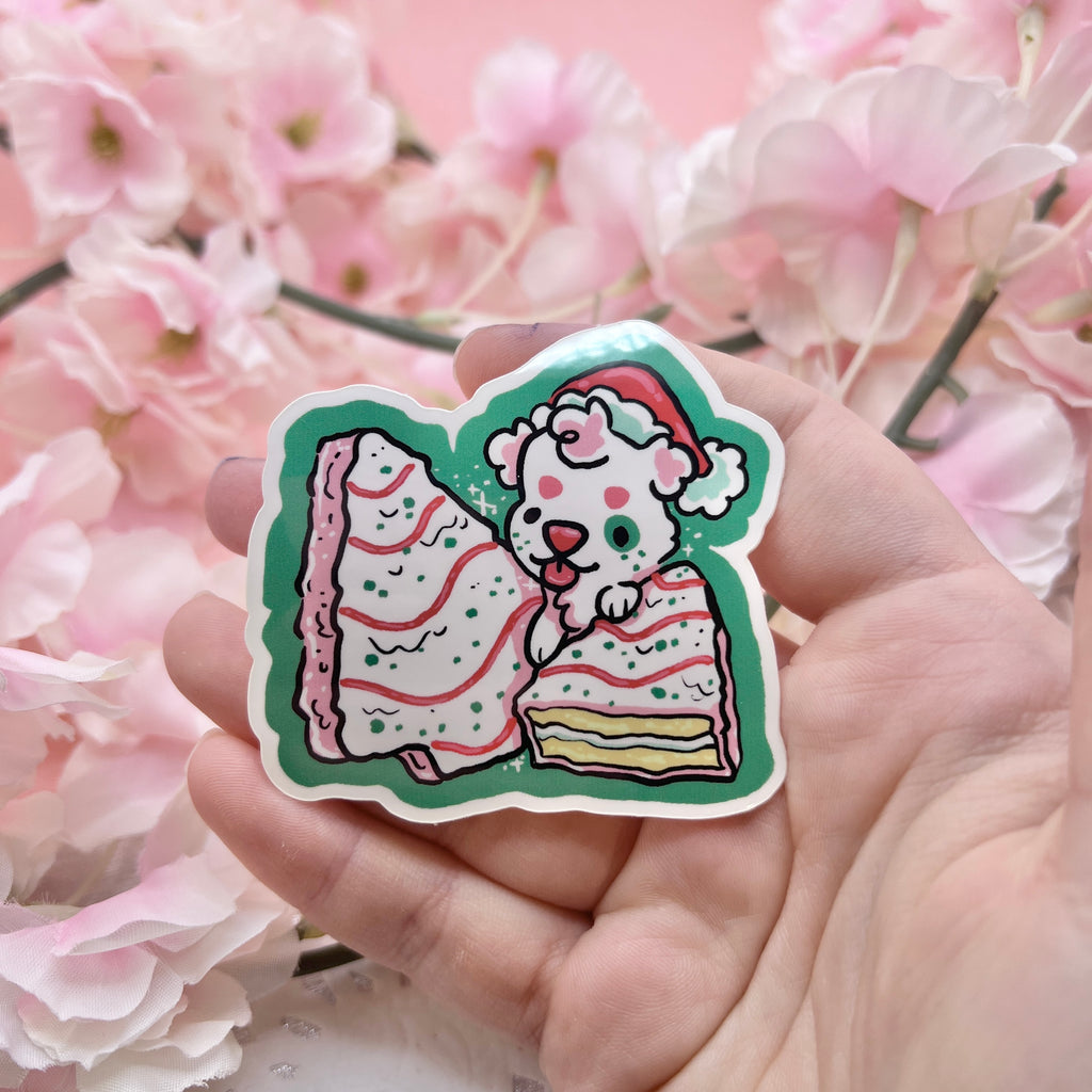 A sticker of a white puppy with a santa hat on top of two white christmas tree snack cakes