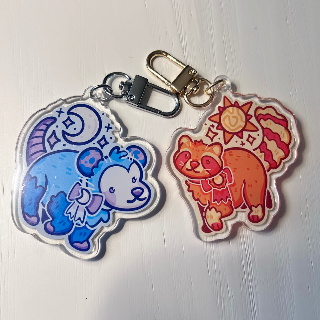 Two colorful keychains featuring cartoon sun raccoon and moon opossum against a white background.
