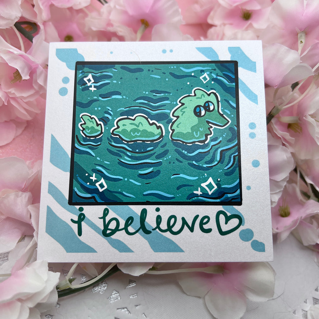 A print that looks like a camera photo, hand written text at the bottom that says "i believe <3", in the photo is lake water with a fuzzy worm lochness monster