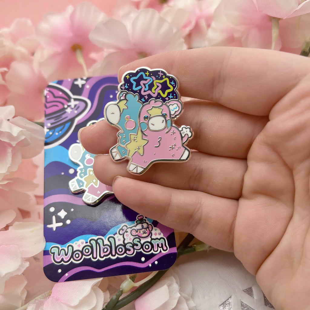 An enamel pin of a two headed calf, one is blue and one is pink, with a yellow star pattern connecting them and on each forehead. On top is a starry sky with three interlocking stars, one blue, one pink, and one yellow.