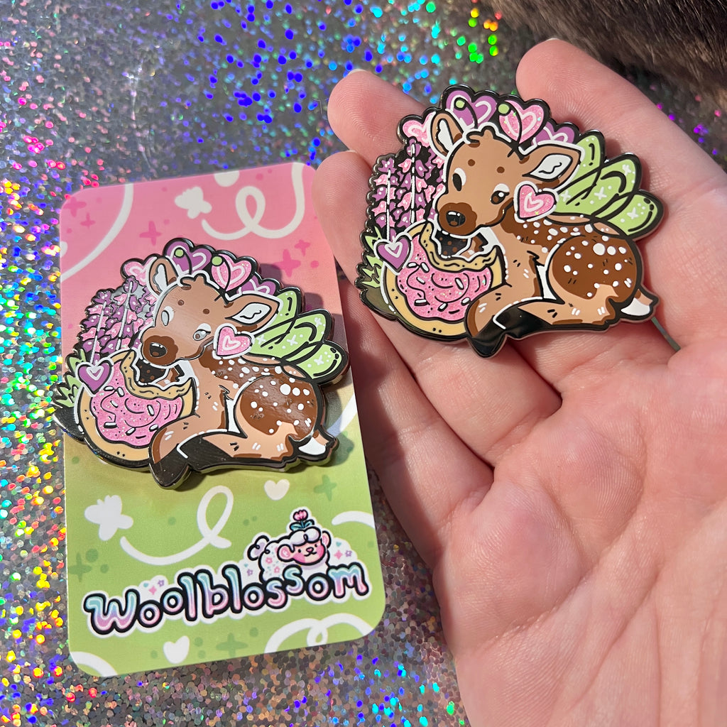 An enamel pin of a fairy deer eating a sugar cookie held by a hand, displayed on a glittery background.