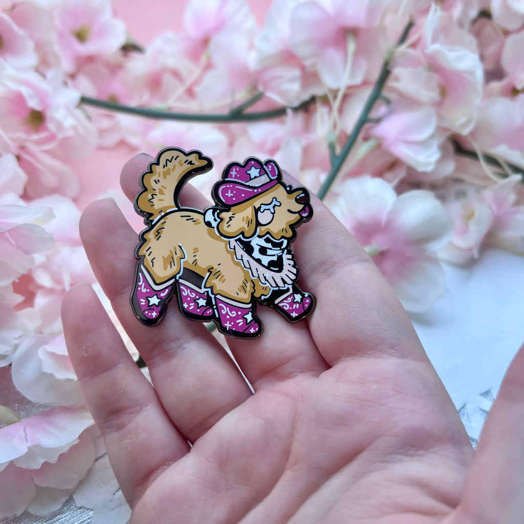 An enamel pin of a golden retriever happily stepping, dressed in a pink cowgirl hat, boots, and bandanna 