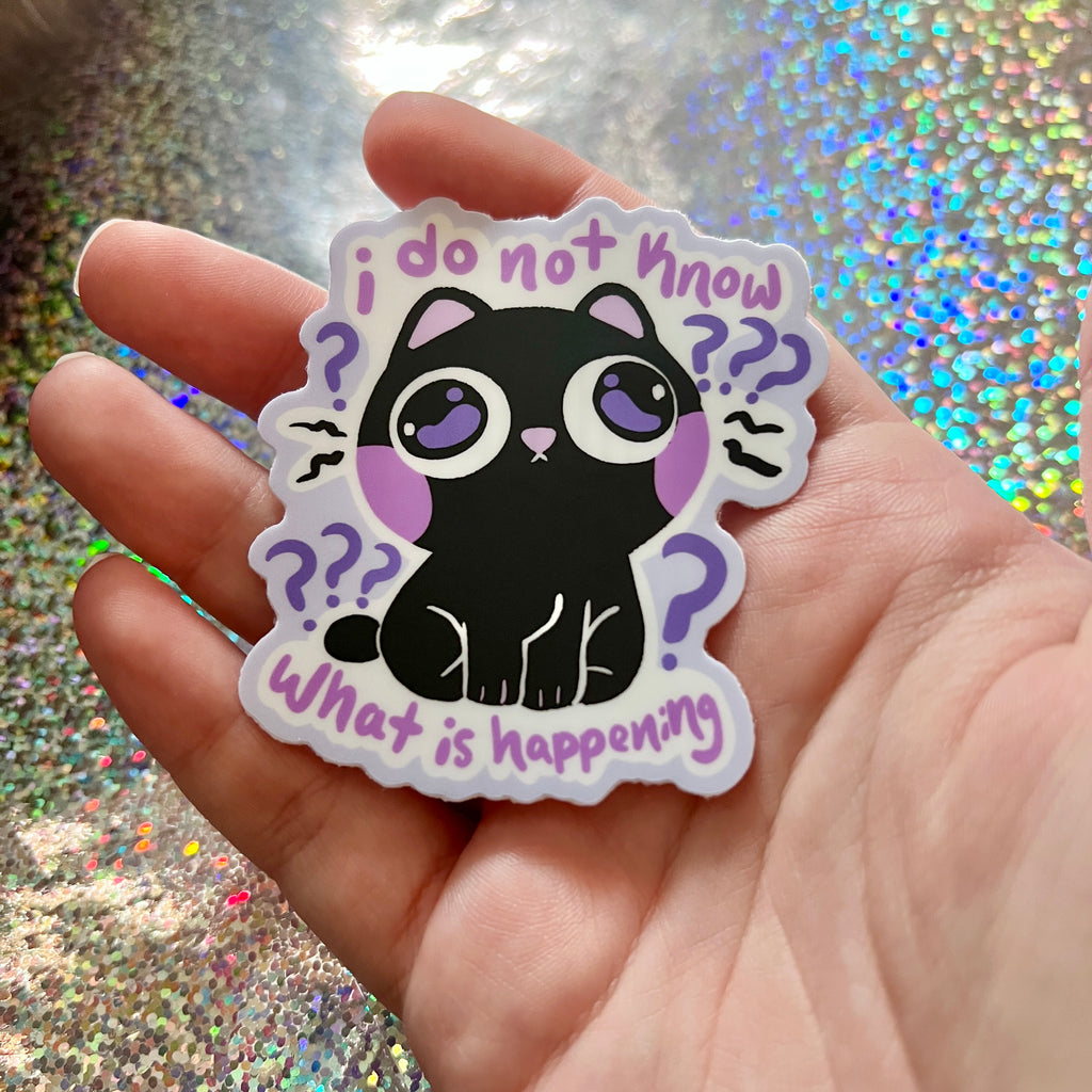 Sticker with a black cat illustration that says "i do not know what is happening" held in a hand against a glittery background