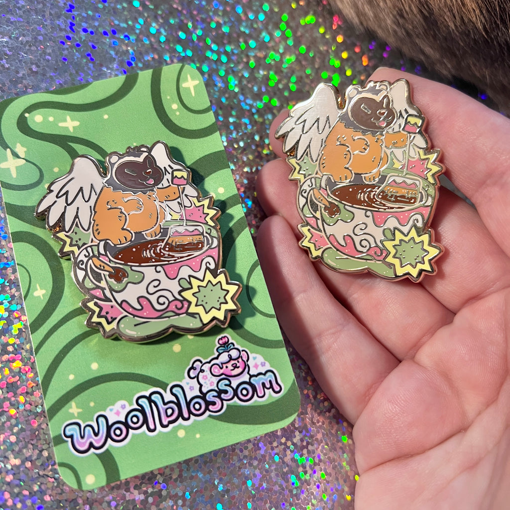An enamel pin of a badger gryffin next to a teacup held by a hand with a glittery background, featuring the brand 'woolblossom'.
