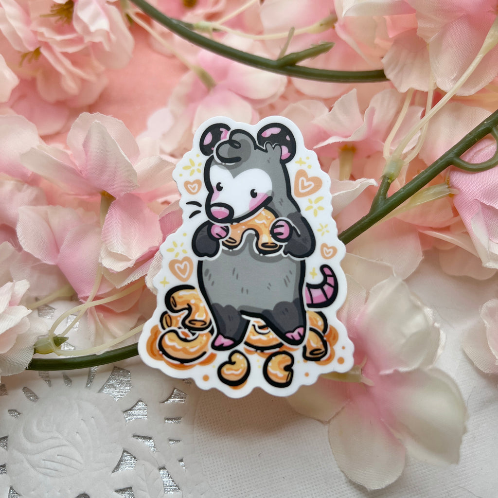 A sticker of an opossum standing in front of a pile of mac and cheese, holding one in its arms up to its mouth with hearts