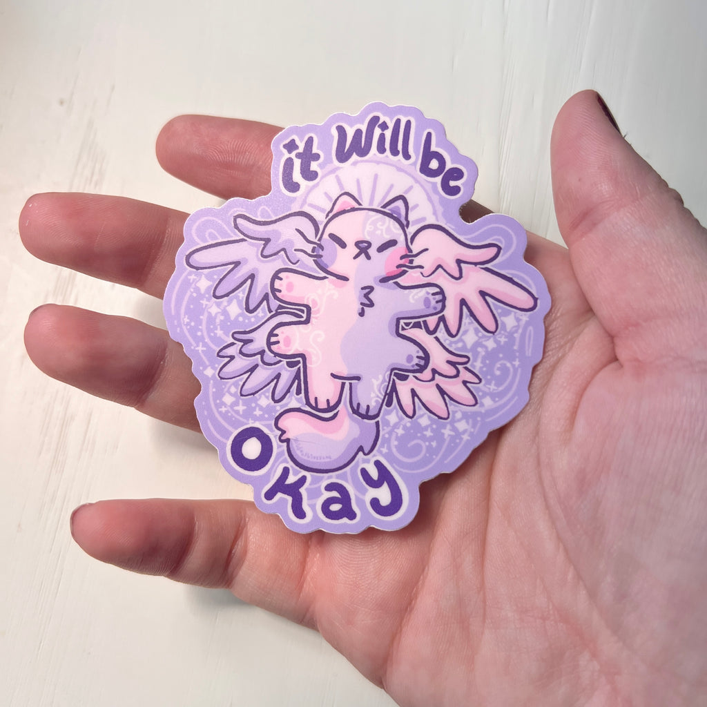 Sticker with a cartoon angel cat and 'it will be okay' text held by hand