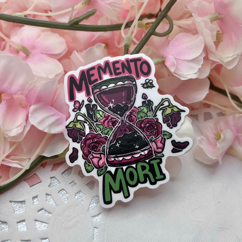 A sticker of a red and black hour glass surrounded by roses at various stages of their lifespan with text that says “memento mori”