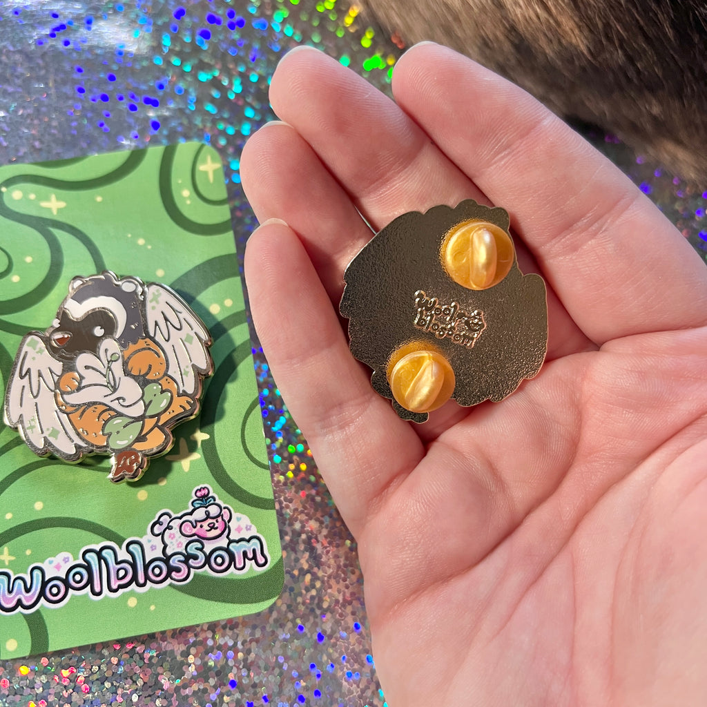 Hand holding an enamel pin of a badger gryffin holding a honeysuckle with a green card in the background.