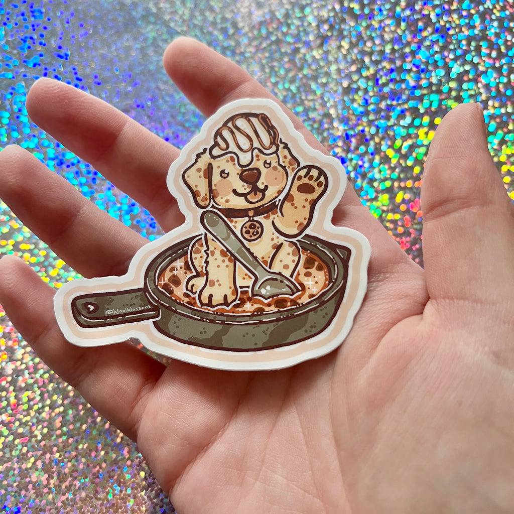 Sticker of a cookie dog on a cookie skillet in a hand against a glittery background