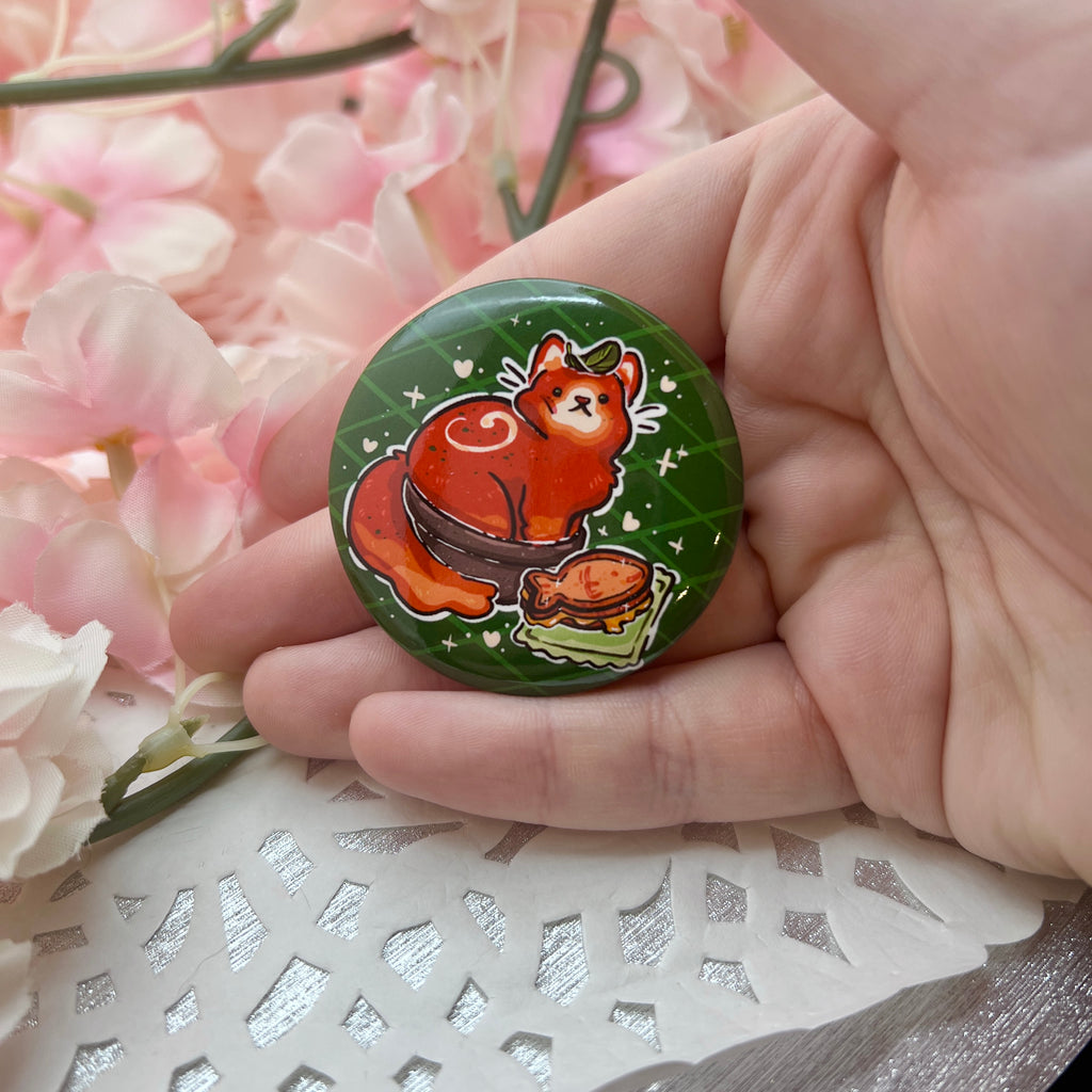 A button of a red tomato soup themed cat in a bowl, basil leaf on head, with a fish grilled cheese next to them.