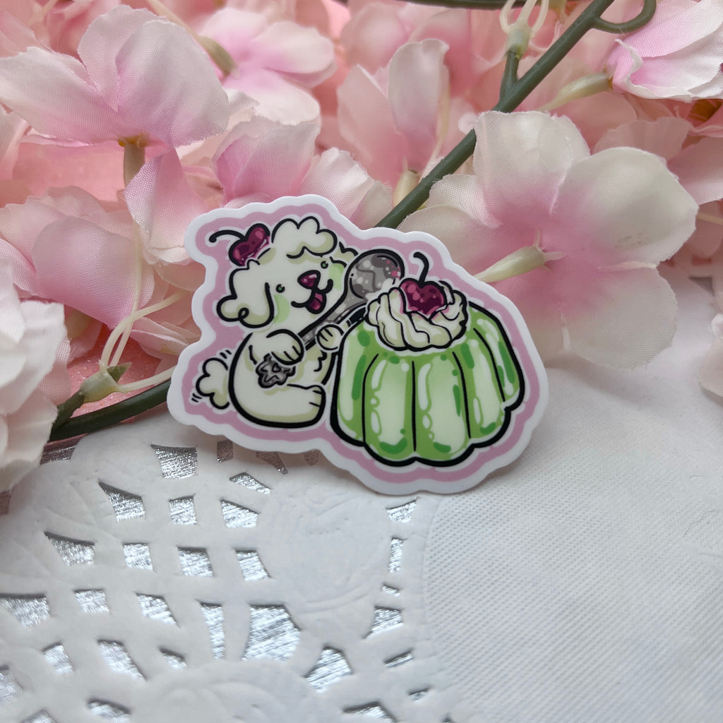 A sticker of a white puppy with a cherry on its head holding a spoon, about to dig into like green gelatin