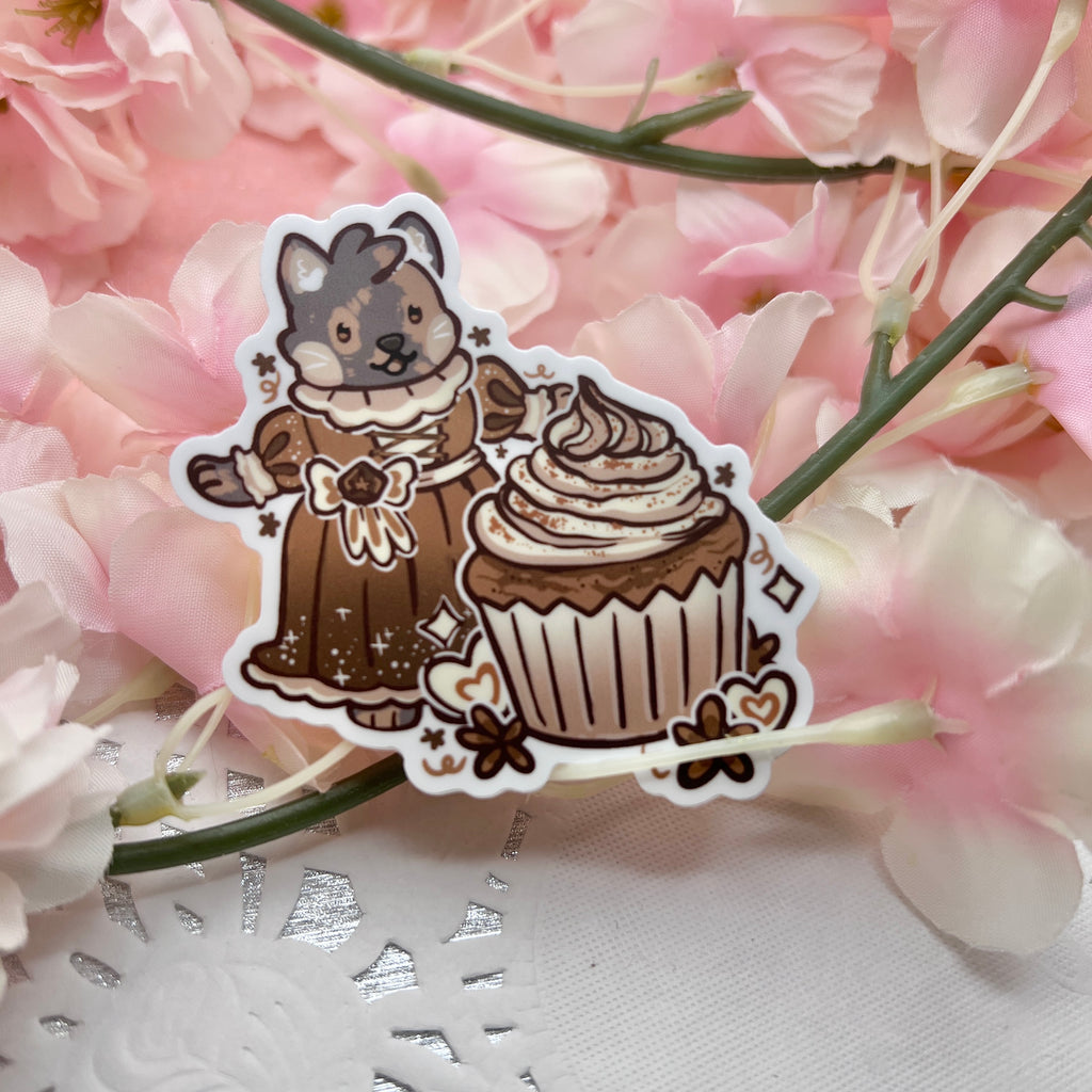 A sticker of my cat Chai dressed in a renaissance/victorian dress, in front of a chai cupcake, and anisse themed