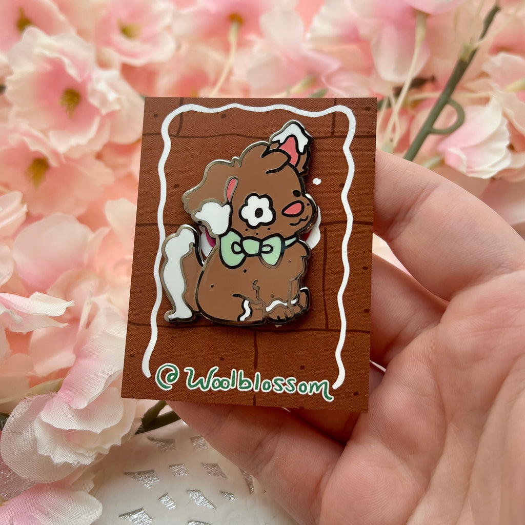 A black nickel enamel pin of Clove the gingerbread puppy, a brown puppy with white frosting accents on her ears and tail, as well as green bow.