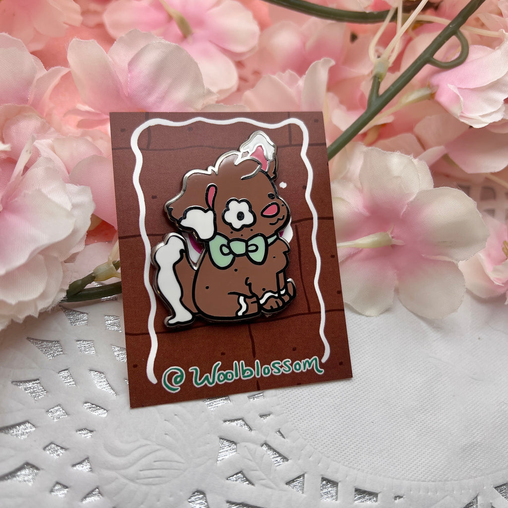 A black nickel enamel pin of Clove the gingerbread puppy, a brown puppy with white frosting accents on her ears and tail, as well as green bow.