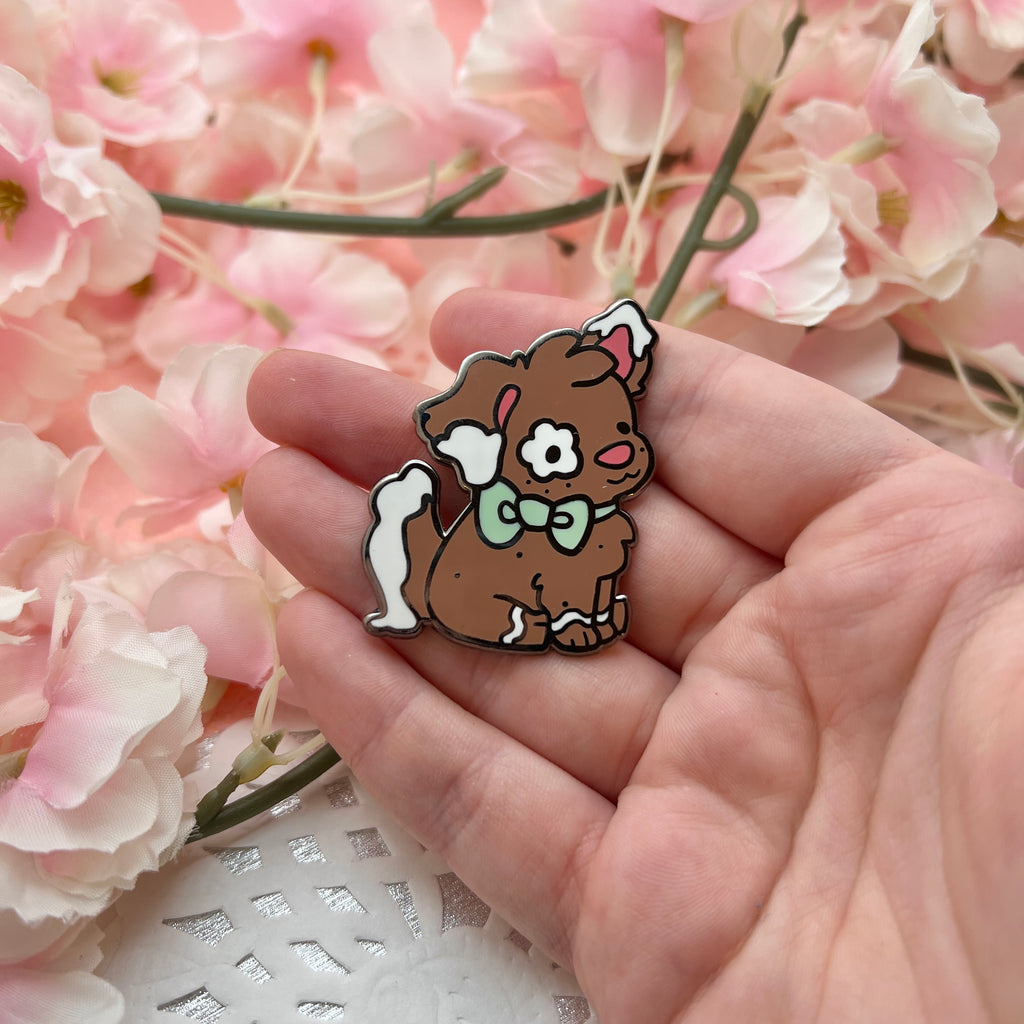 A black nickel enamel pin of Clove the gingerbread puppy, a brown puppy with white frosting accents on her ears and tail, as well as green bow.