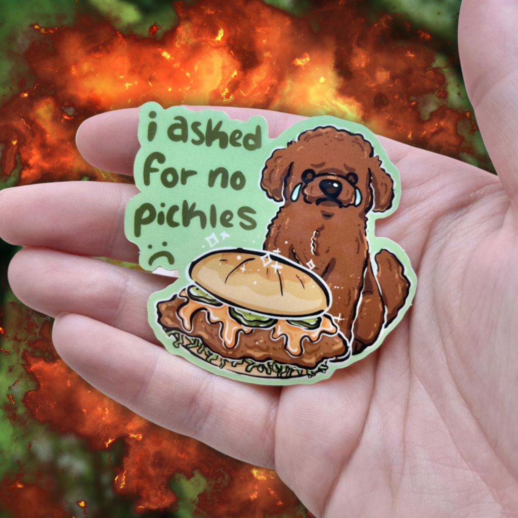 A sticker of a brown poodle dog sad and crying in front of a chicken sandwich that has pickles. Text says "i asked for no pickles :(" against a green background