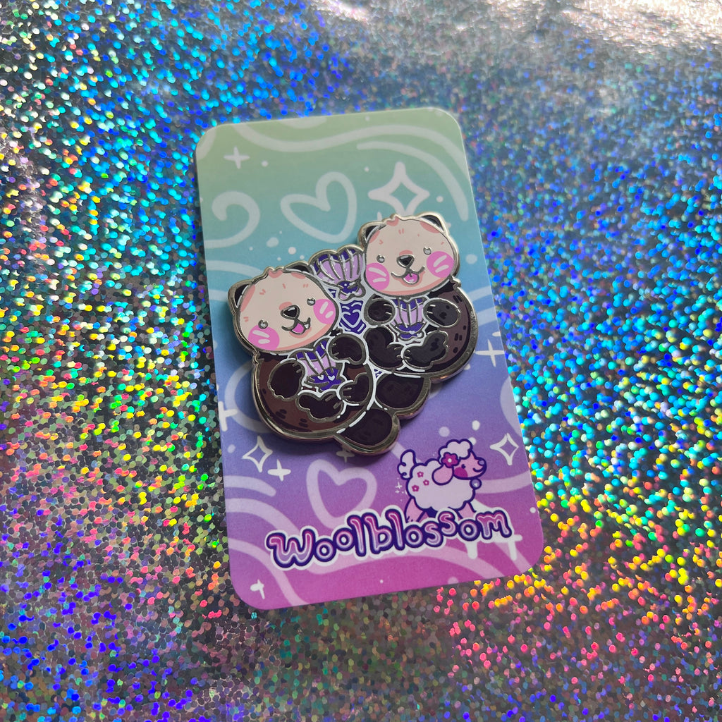 An enamel pin of two otters floating together holding heart shaped seashells