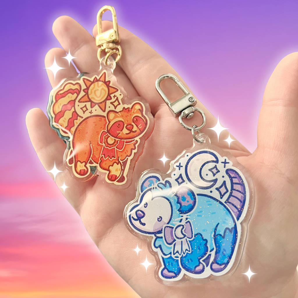 Two colorful keychains featuring cartoon sun raccoon and moon opossum held in a hand against a gradient background.