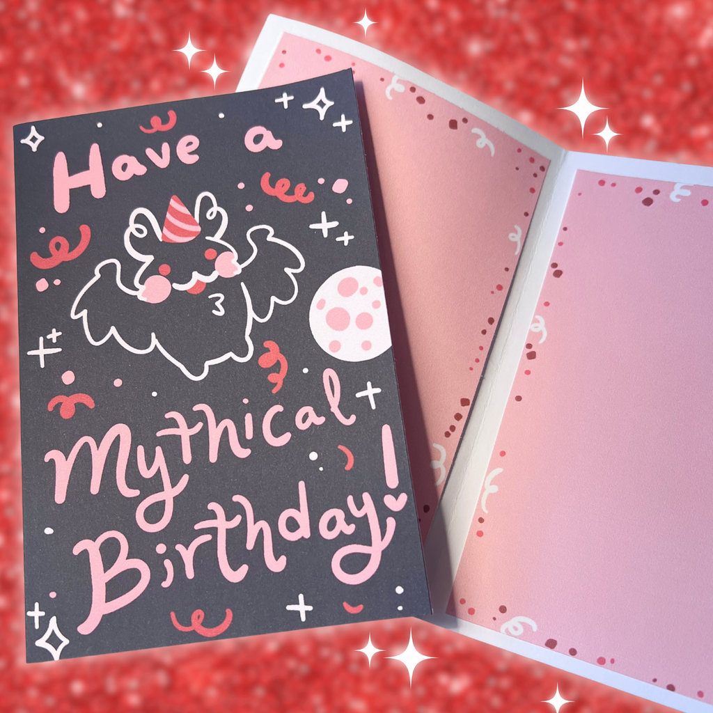 Hand holding a card with a mothman wearing a birthday hat and text 'Have a mythical birthday!' on a red background.