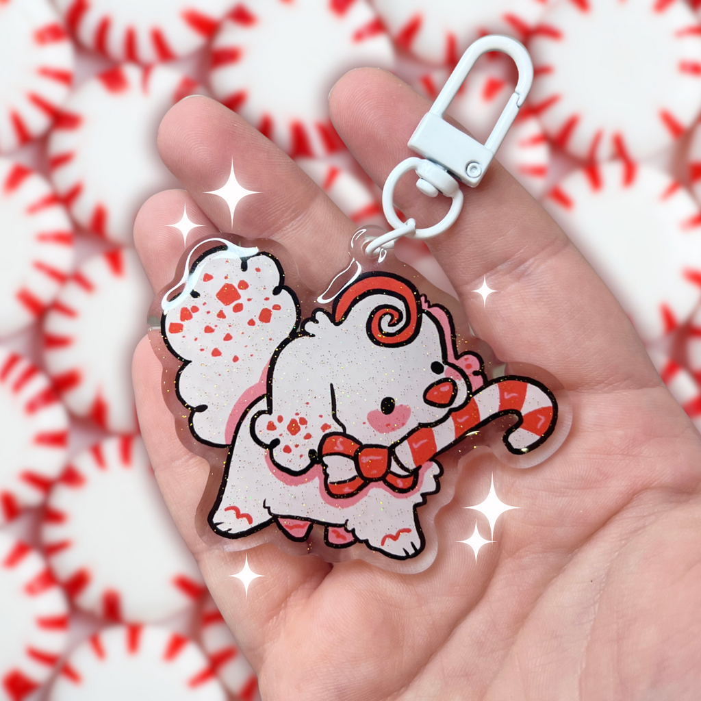 A keychain charm of a white peppermint bark themed puppy holding a candy cane in her mouth