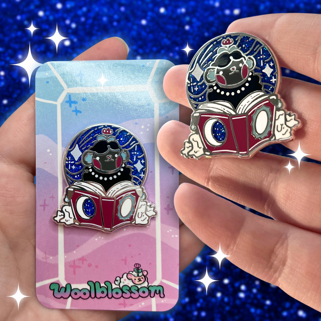 Pin with a design of a sheep reading a book held by a hand, with 'Woolblossom' branding on a blue starry background.