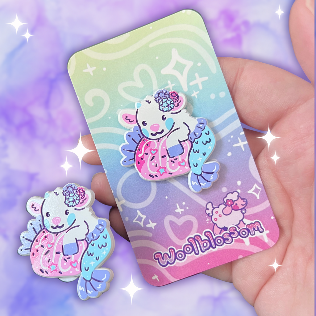 Enamel pin of a mer-goat holding a strawberry on a card with 'woolbbloom' branding against a purple and pink gradient background.