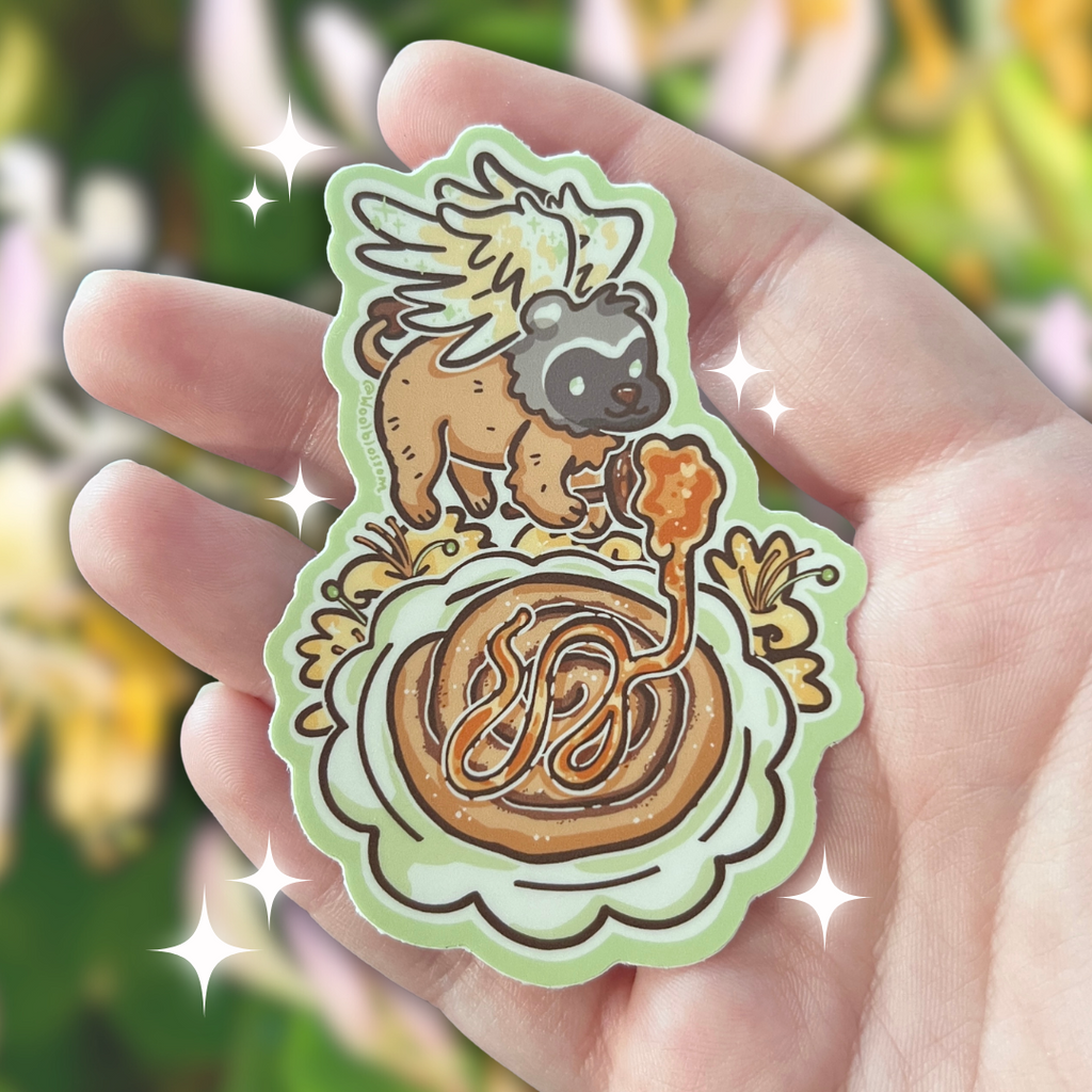 Hand holding a sticker of a badger gryffin pouring honey over a cinnamon roll with honeysuckles against a blurred natural background