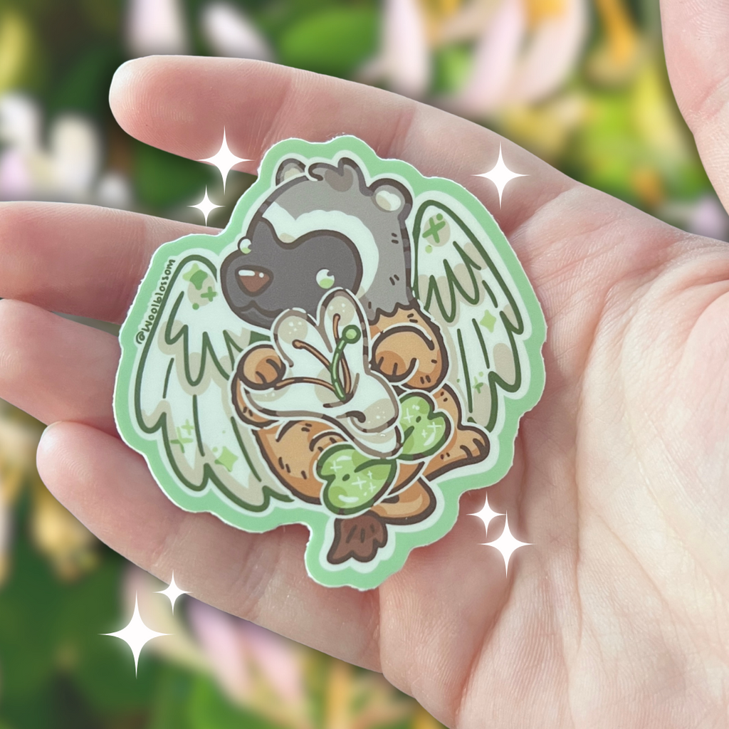 Hand holding a sticker of a badger gryffin holding a honeysuckle against a blurred natural background