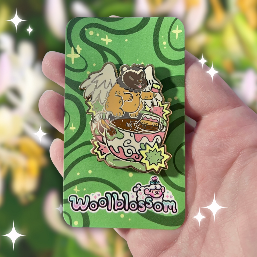 An enamel pin of a badger gryffin next to a teacup on a card with 'woolblossom' branding held in a hand against a blurred floral background.