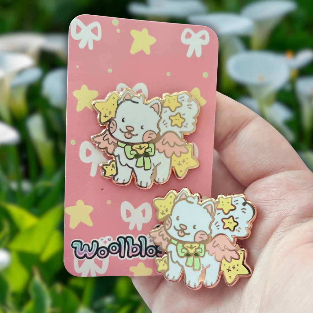 An enamel pin of a white cat with pink wings and yellow stars on a pink card with 'Woolblossom' branding, held against a blurred green leafy background.