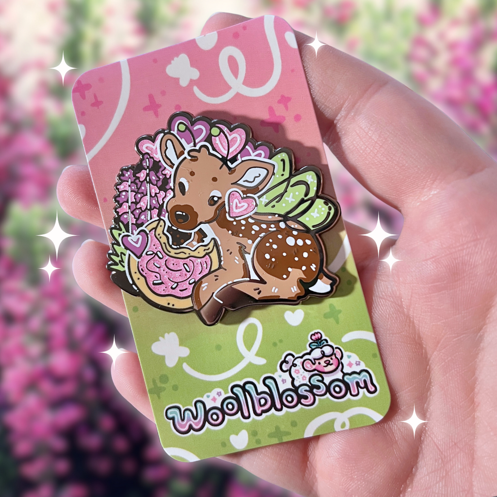 An enamel pin of a fairy deer eating a sugar cookie on a colorful card held by a hand with a floral background
