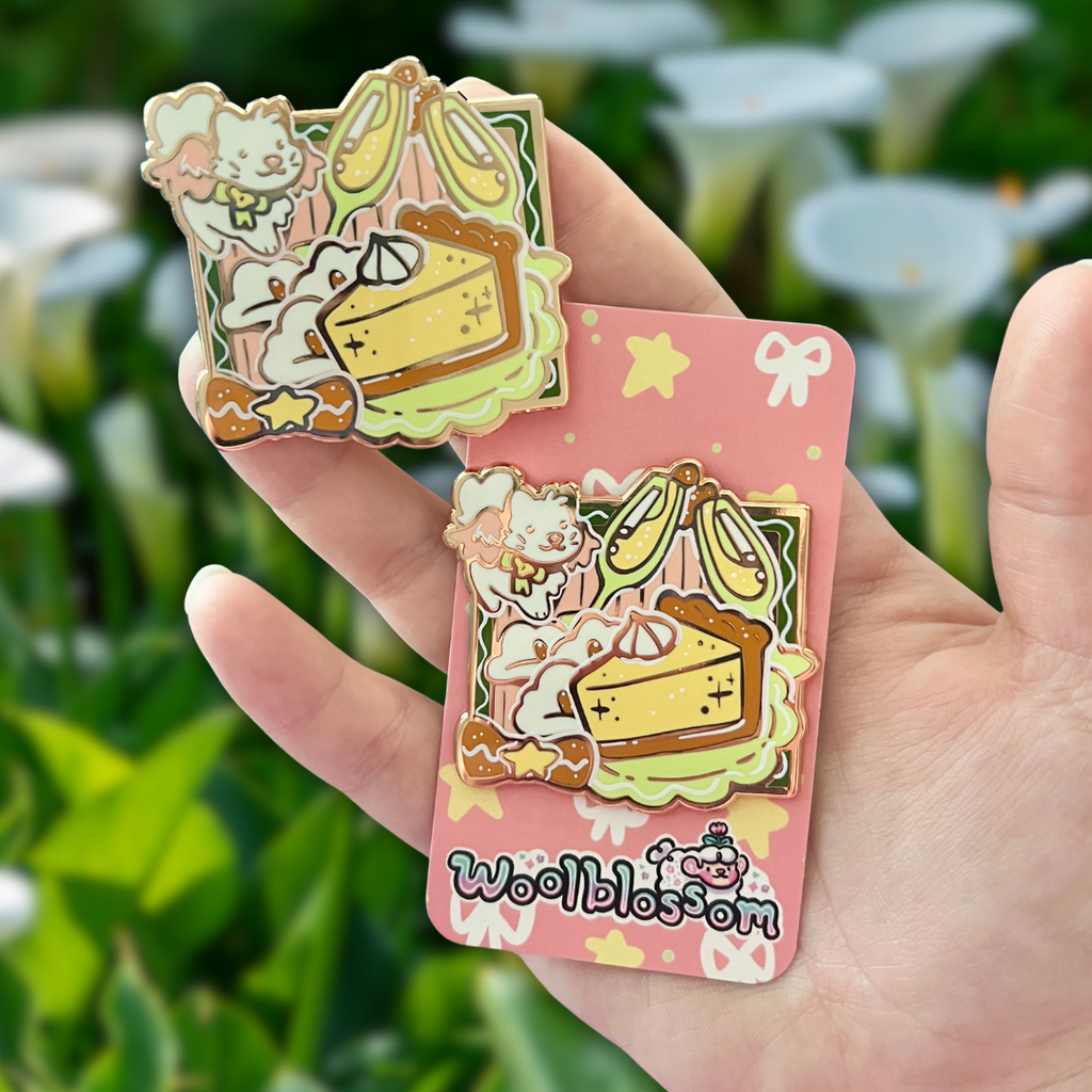Hand holding an enamel pin of a white angel cat flying around a celebration meal  pink card with 'woolblossom' branding against a blurred green background.