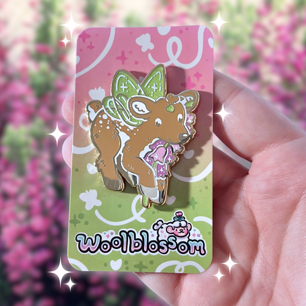 An enamel pin of a fairy deer holding a heather flower on a 'woolblossom' branded card, held against a blurred floral background.