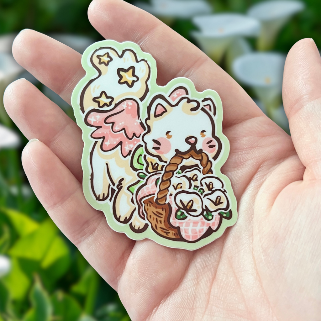 Hand holding a cute sticker of a cat with angel wings and a basket of flowers against a blurred green background.