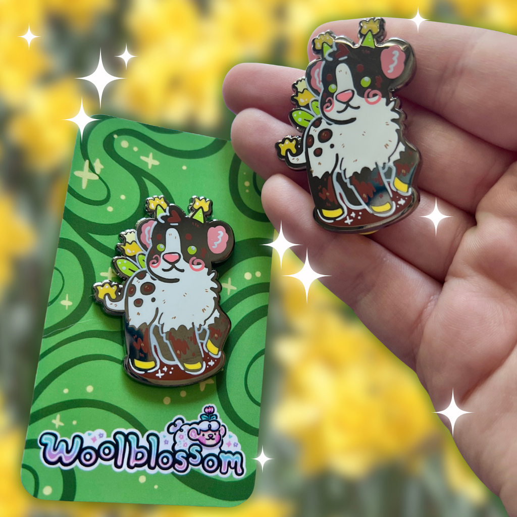 Cartoon alien cow pin held by a hand with a green card displaying the brand 'Wool Blossom' against a yellow floral background.