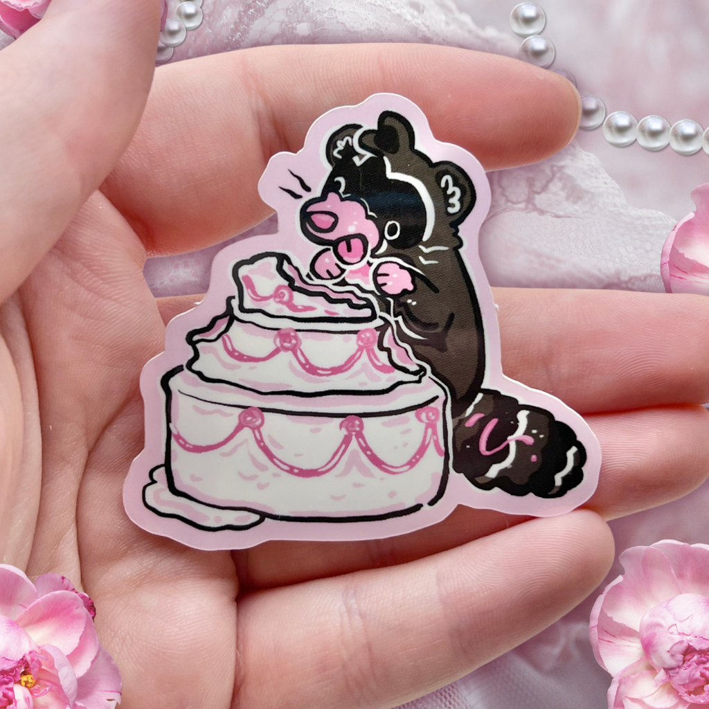 Sticker of a raccoon eating cake held in a hand with a floral background