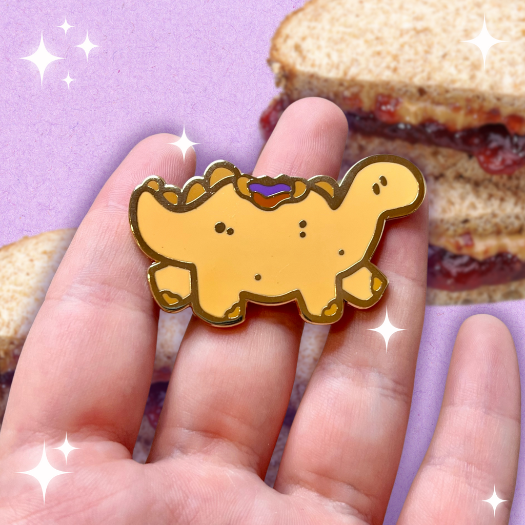 A gold enamel pin of a tan colored stegosaurus inspired dino that resembles a PBJ sandwich, the crustless kind, with a bite taken out of its back so you see the PB and jelly