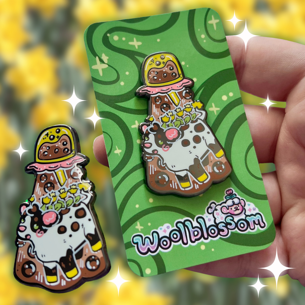 An alien boba cow being abducted by a boba enamel pin on a green card with 'Woolblossom' branding, held against a blurred yellow background.