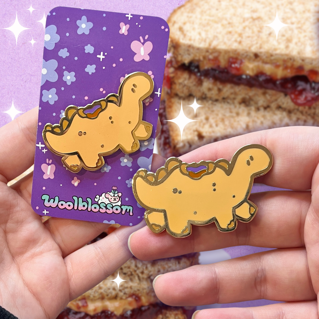 A gold enamel pin of a tan colored stegosaurus inspired dino that resembles a PBJ sandwich, the crustless kind, with a bite taken out of its back so you see the PB and jelly