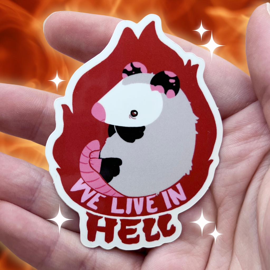 Sticker with cartoon character opossum and 'We Live in Hell' text held in a hand against a fiery background
