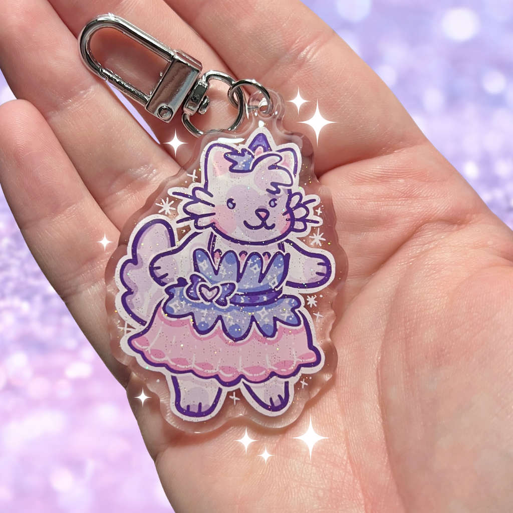 keychain of a cat in a pink dress held in a hand with a purple glittery background